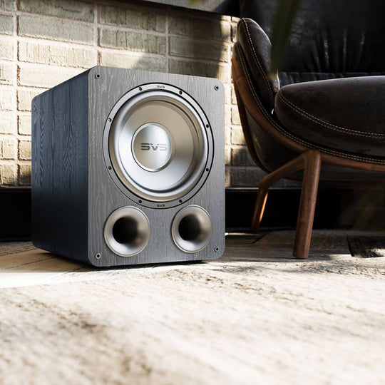 The SVS PB-1000 Pro - Ported Box Home Subwoofer in Black Ash from Todds Hi Fi resting on the floor beside a leather and dark wooden statement chair