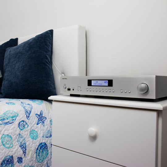 The Rotel A14 MKII Stereo Integrated Amplifier in Silver from Todds Hi Fi sitting on a white wooden bedside table next to a bed with beachy bedding and dark blue statement cushions