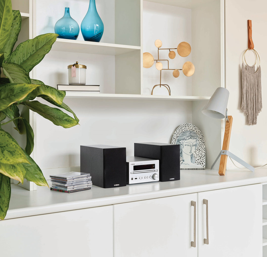 The Yamaha MCRB270D Micro HIFI in silver from Todds Hi Fi placed on a cabinetry unit with over-head open storage, cd's and a table lamp next to it